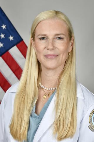 Blonde woman in white coat with US flag behind her.