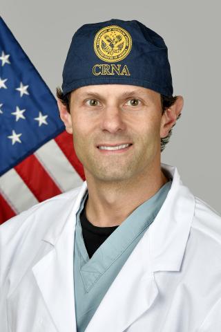 Man in white lab coat and blue cap standing in front of an American flag.
