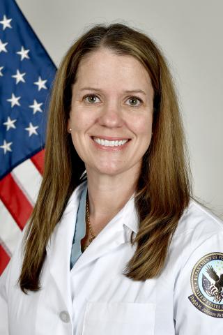 A smiling woman in a white coat stands in front of an American flag.