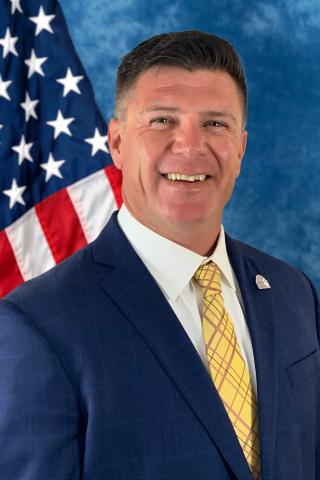 Man in a blue suit and yellow tie smiling in front of an American flag.