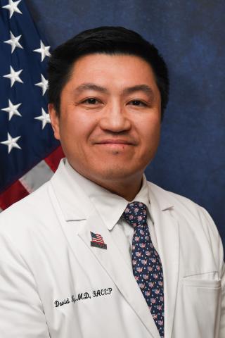 Smiling man in white coat and tie with American flag behind him.