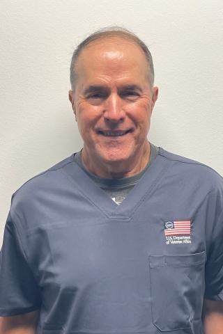 Smiling man in navy scrubs with US flag and text on chest pocket.