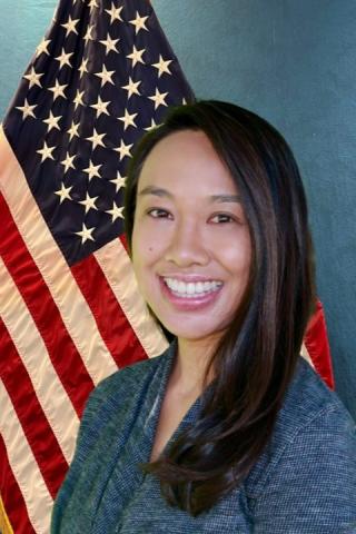 A smiling woman with long black hair stands in front of an American flag.