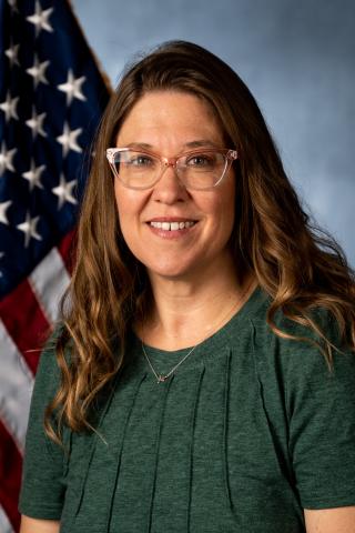 A smiling woman with long hair in a green blouse stands in front of an American flag.