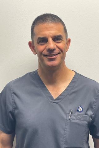 Middle-aged man in gray scrubs smiling against a plain white wall.