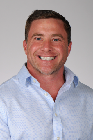 Smiling man in blue shirt with rolled up sleeves against plain white background.