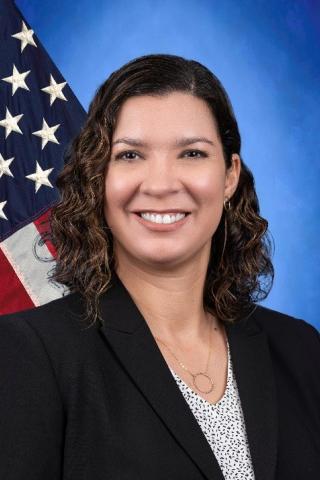 Woman in a black blazer and white top, smiling in front of an American flag.