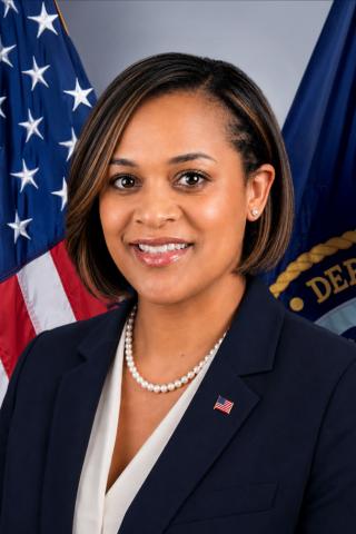 Portrait of a smiling woman in a navy blazer with pearl necklace, standing in front of flags.