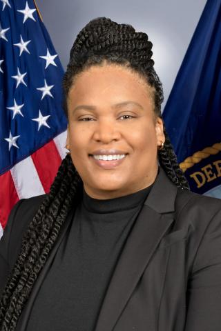 Woman in black blazer smiles in front of USA and Department flag