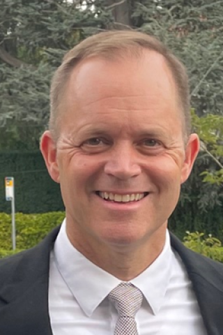 Man in suit and tie smiling in front of green bushes.
