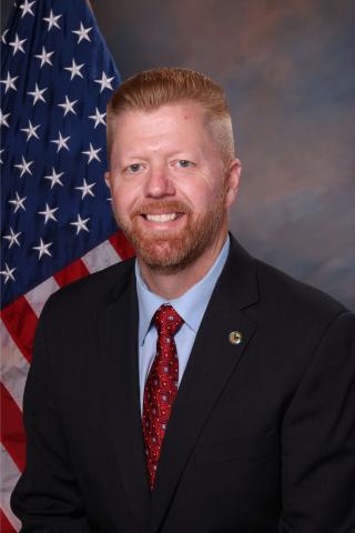 Man in suit and red tie standing before American flag.