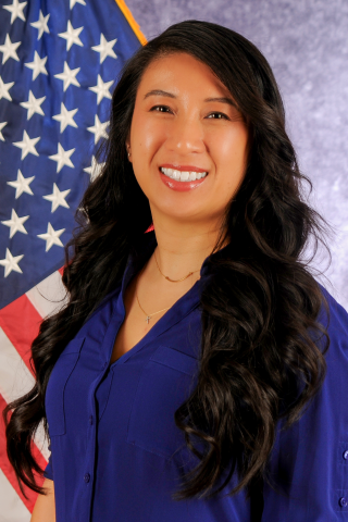 Woman with long dark hair wearing blue shirt smiling in front of American flag.