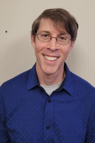 Smiling man in blue shirt and glasses stands against plain background.
