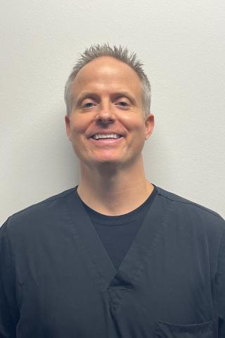 A smiling man in black scrubs stands against a white wall.