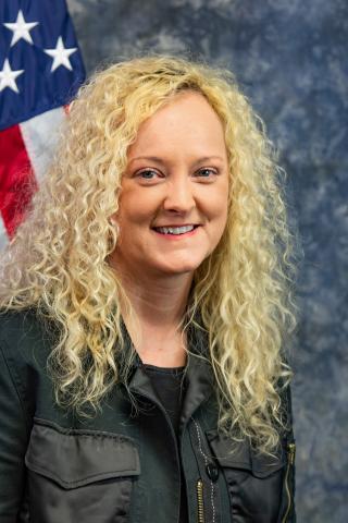 Woman with blonde hair and black jacket standing in front of an American flag.