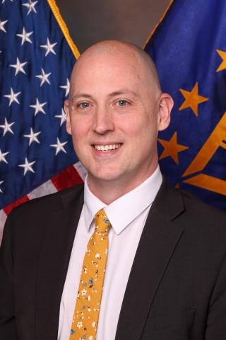 Bald man in black suit and yellow tie smiles in front of American and SES flags.