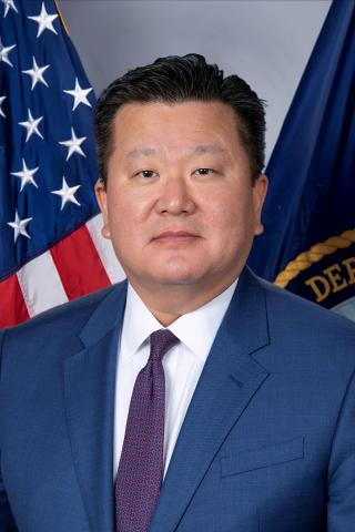 Man in a blue suit and red tie stands in front of American and Department flags.