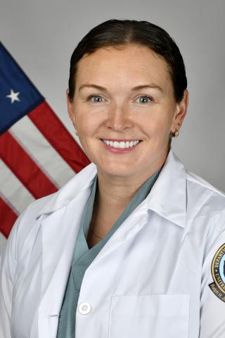 Woman in white lab coat smiling in front of an American flag.
