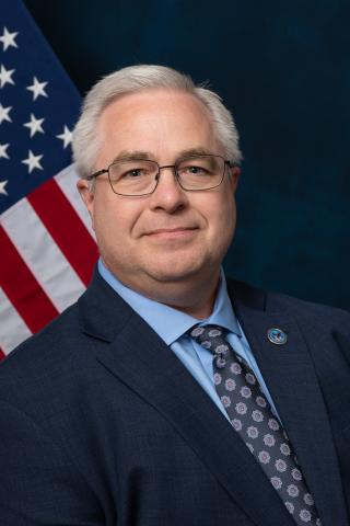 Man in suit and tie smiling in front of American flag.