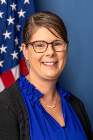 Woman in blue blouse and black cardigan smiling with American flag behind her.