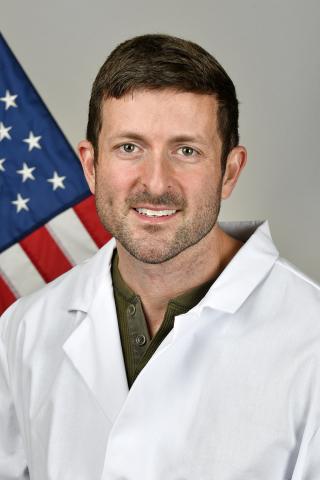 A man in a white lab coat smiles in front of an American flag.