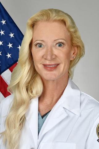 Portrait of a blonde woman in a white lab coat with an American flag in the background.