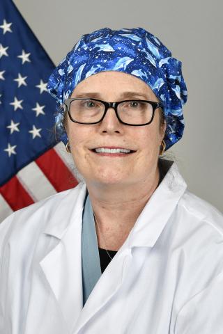 Woman in white lab coat and blue scrub cap smiling in front of American flag.