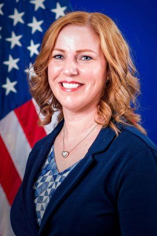 Woman with blonde hair in blue shirt and blazer smiles in front of American flag.