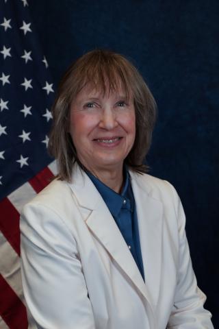 Woman in white lab coat and blue shirt smiling with American flag in background.