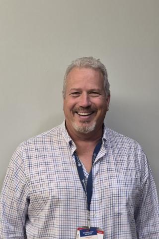 Man with gray hair and beard, wearing blue and white shirt, smiling at camera.