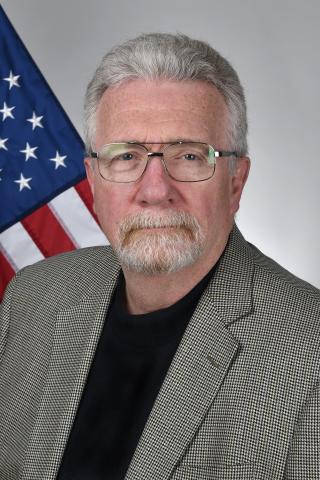 Man with white hair and beard wearing glasses and gray jacket, standing by American flag.