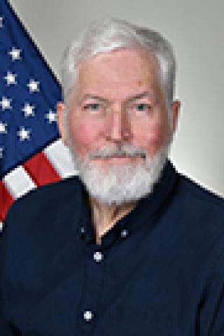 Man with white hair and beard in navy shirt in front of USA flag.