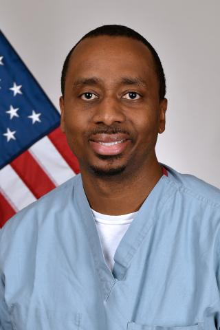 Man in blue scrubs smiling in front of an American flag.