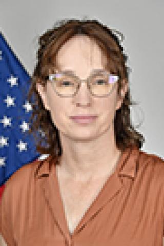 Woman in brown shirt with glasses stands before US flag.