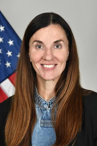 Woman with long brown hair smiling in front of an American flag.
