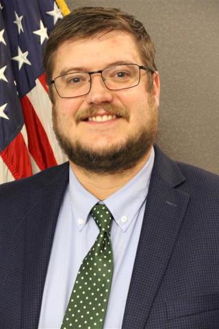 Man in a suit and green tie smiling in front of an American flag.