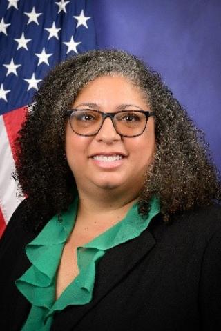 Woman in black blazer and green shirt smiling with American flag in background.
