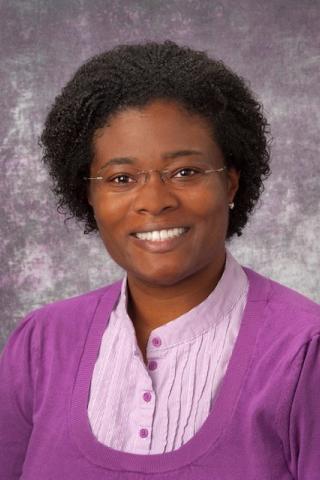 Woman with short curly hair wearing glasses, a purple cardigan, and pink shirt smiling.