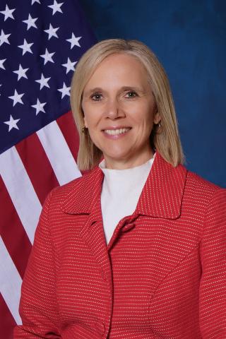 Blonde woman in red blazer and white shirt smiles in front of US flag.