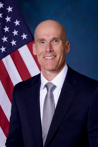 Man in suit and tie smiling with American flag in the background.