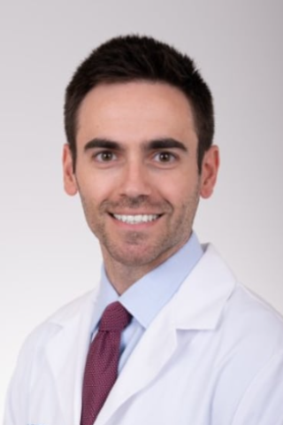 Man in white lab coat and maroon tie smiling at camera.