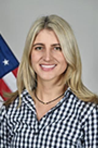 Woman with blonde hair and checkered shirt smiling with American flag behind her.