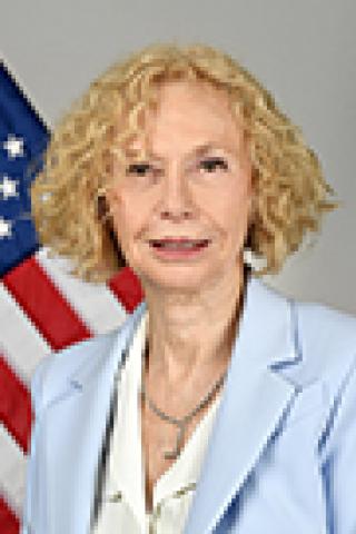 Blonde woman in blue blazer and white blouse with American flag in background.