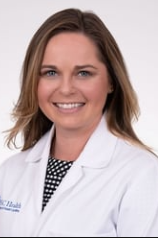 Female doctor with brown hair, smiling, wearing white lab coat with blue logo.