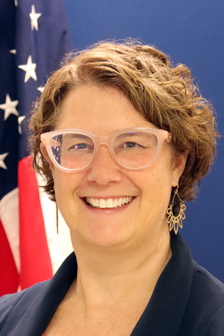 Woman with short hair, glasses, and navy blazer smiling with US flag in background.