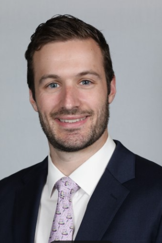 Man in suit and purple tie smiling against a gray background.