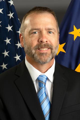 Man in a suit and tie smiling with USA and EU flags behind him.