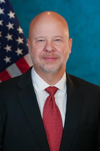 Smiling bald man in black suit, red tie, and white shirt with American flag behind.