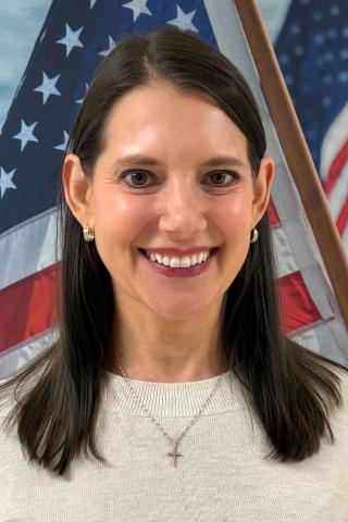 Woman with long black hair and light sweater, smiling in front of American flag.