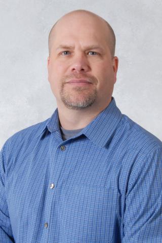 Man with bald head and blue shirt standing against a plain background.